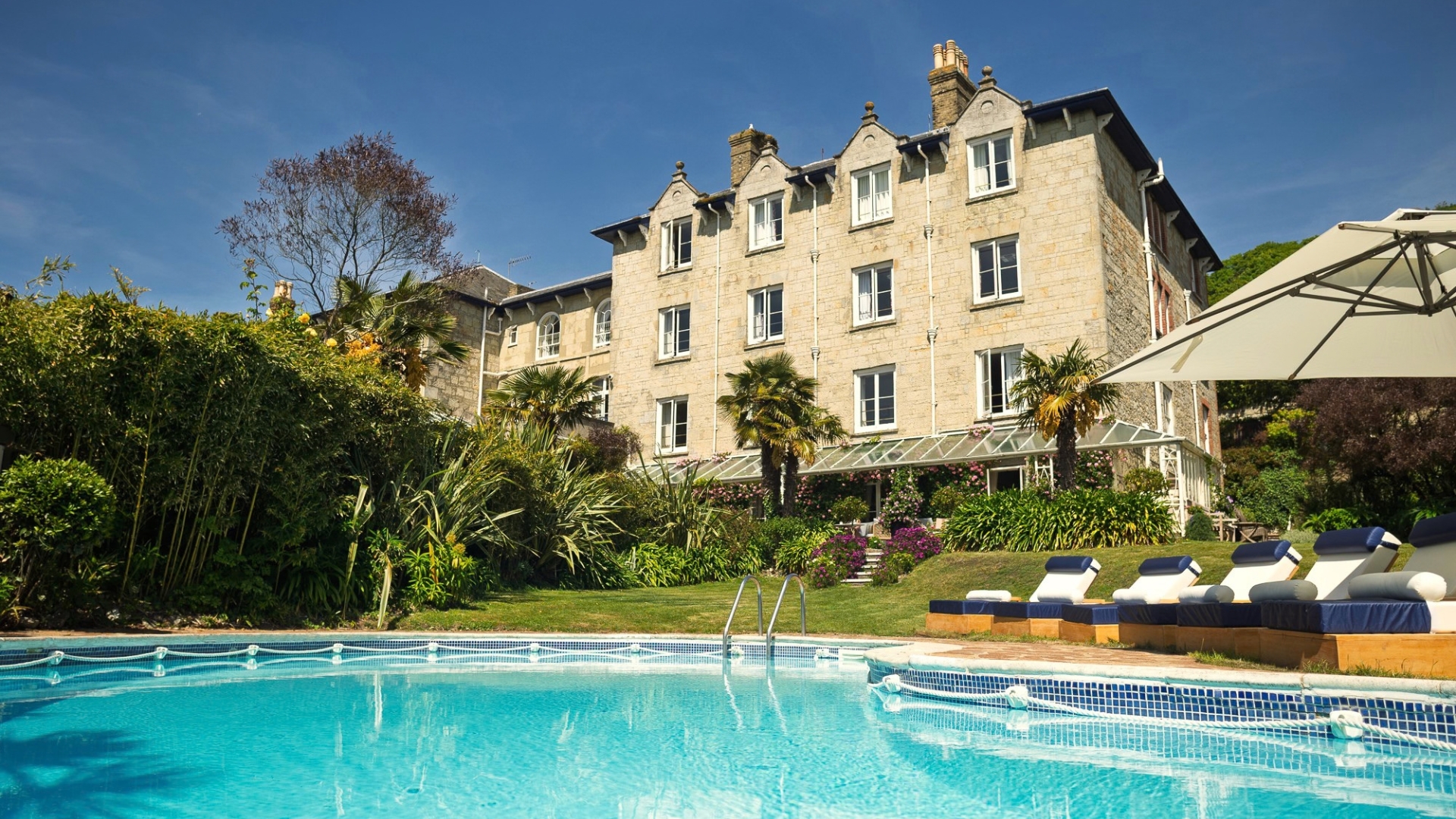 Outdoor swimming pool surrounded by sun loungers, landscaped gardens and palm trees, with The Royal Hotel's historic stone building rising behind. Clear blue skies and well-maintained grounds create a relaxing holiday atmosphere.