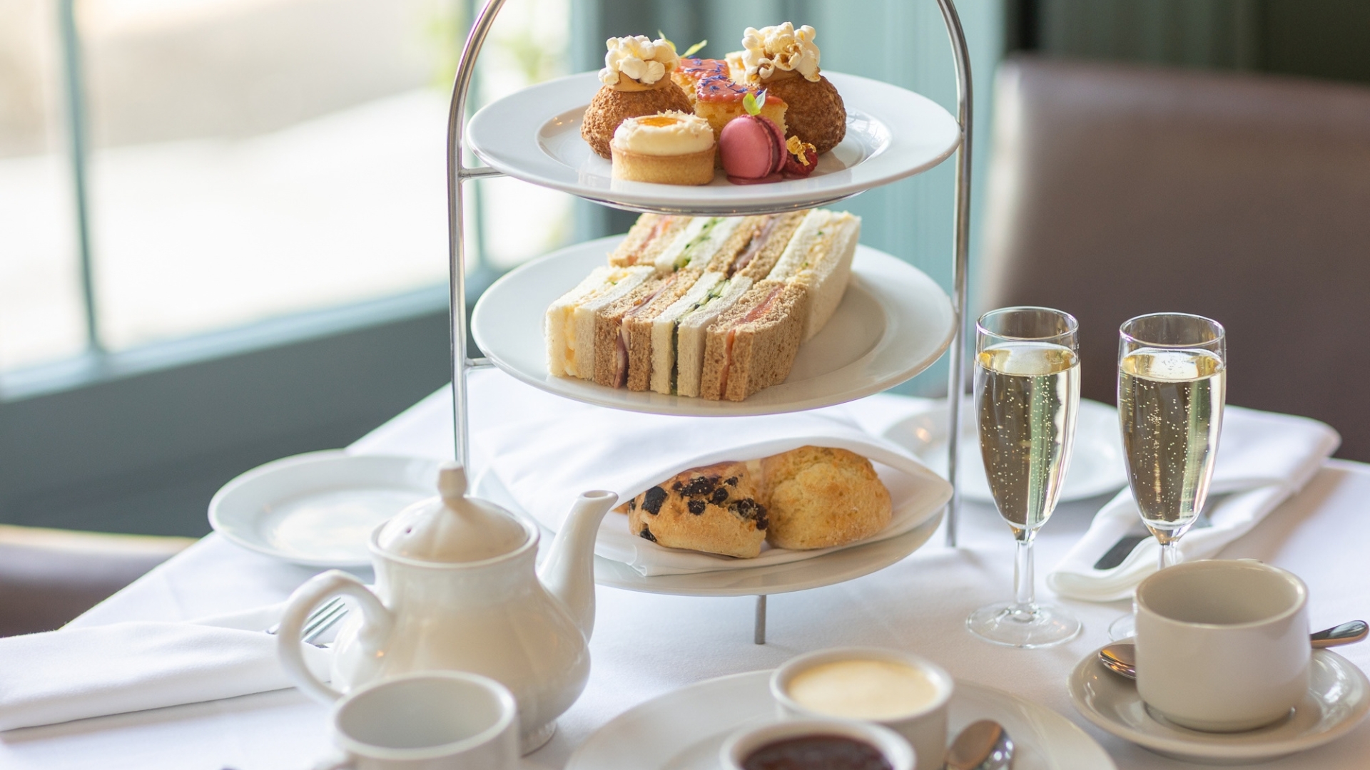 Traditional afternoon tea served on a three-tier stand, featuring finger sandwiches, scones and an assortment of cakes and pastries. A teapot, cups and two glasses of sparkling wine are arranged on a white tablecloth, creating an elegant dining experience.
