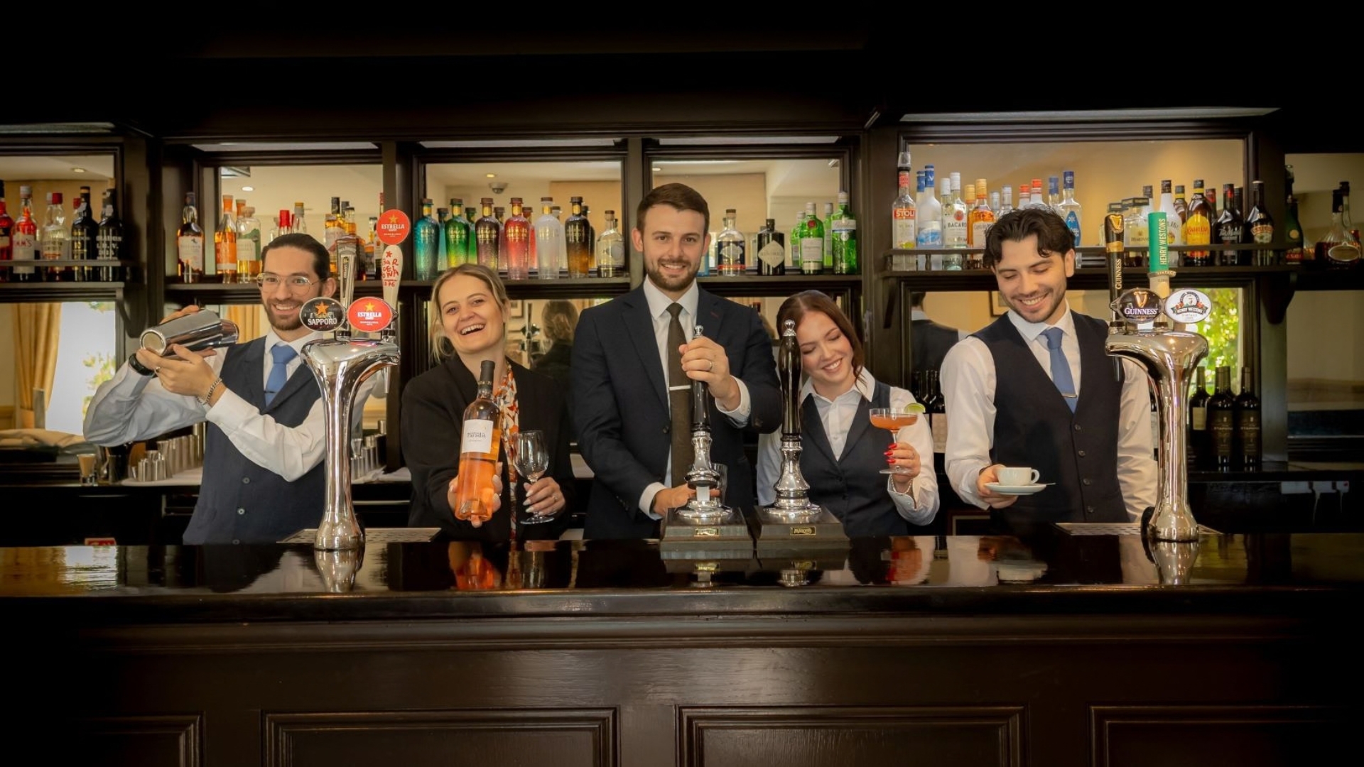Hotel team standing behind a well-stocked bar, preparing and serving a selection of drinks including cocktails, coffee, wine and draught beverages. Shelves of spirits and bottles line the background, highlighting the hotel's hospitality offering.
