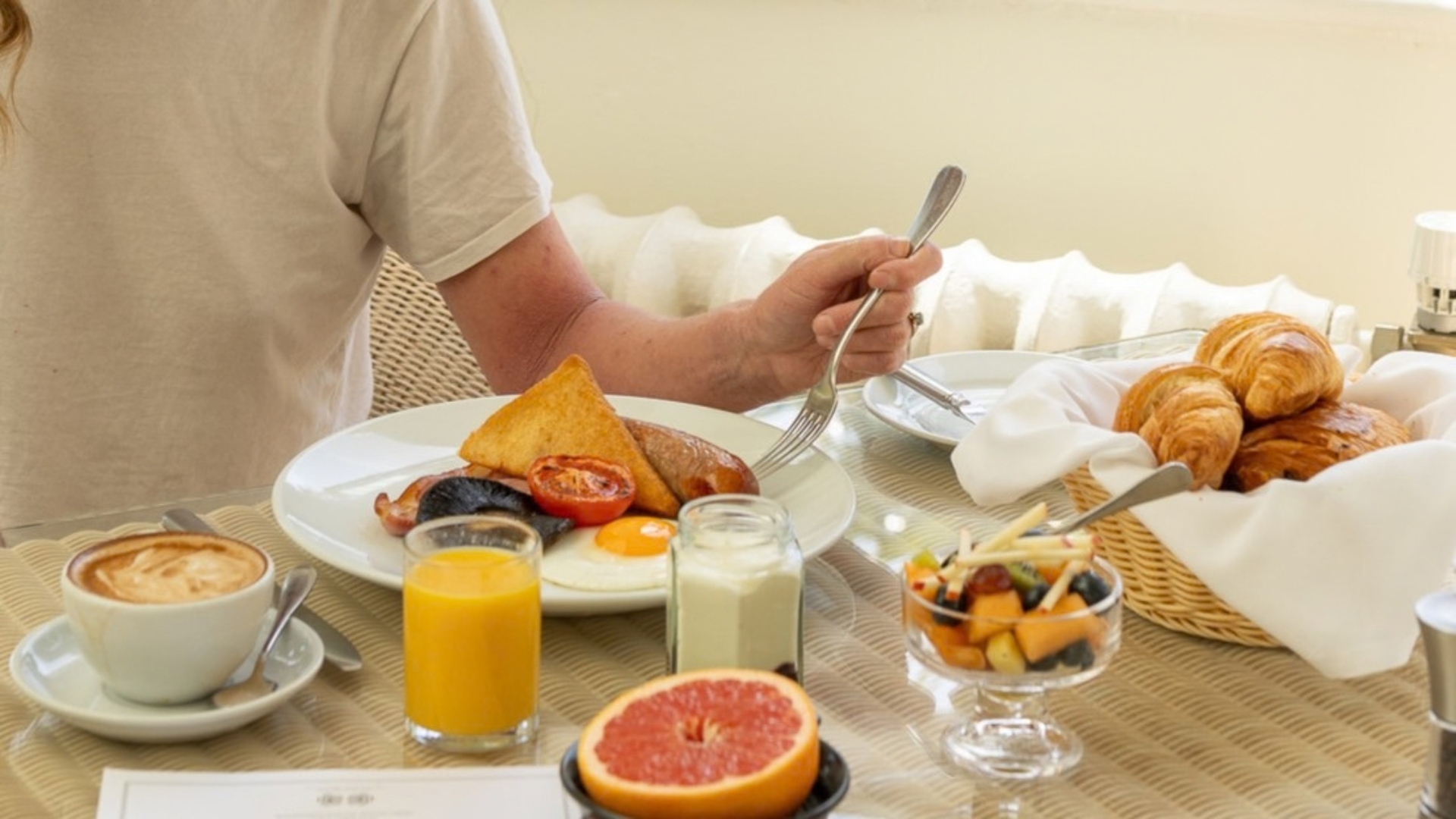 Hotel breakfast spread featuring a cooked breakfast with eggs, sausage, bacon, black pudding and toast, alongside croissants, fresh fruit, yoghurt, orange juice and coffee. The meal is served at a table in a bright dining setting, showcasing a hearty breakfast experience.