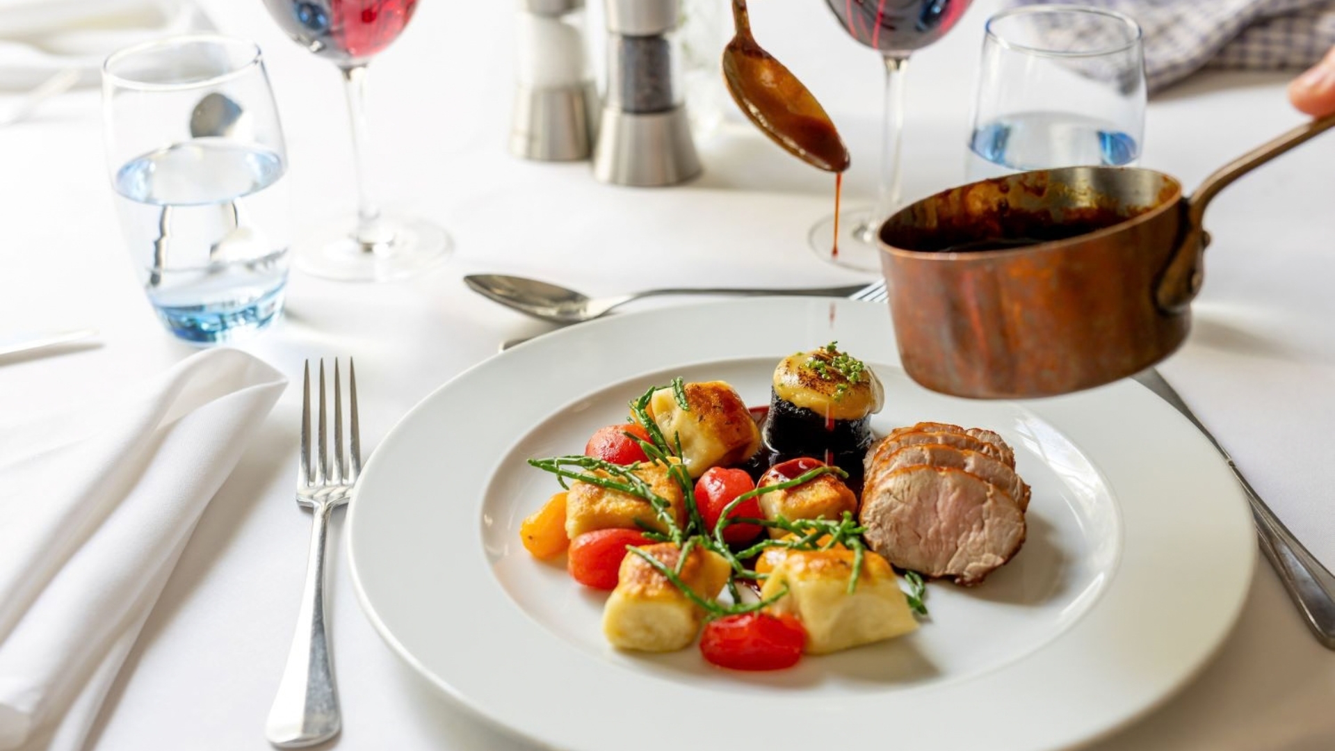 Fine dining dish served on a white tablecloth, featuring sliced meat, roasted vegetables and golden potato accompaniments. A rich sauce is being poured from a copper pan, with wine glasses, water glasses and polished tableware creating an elegant restaurant setting.