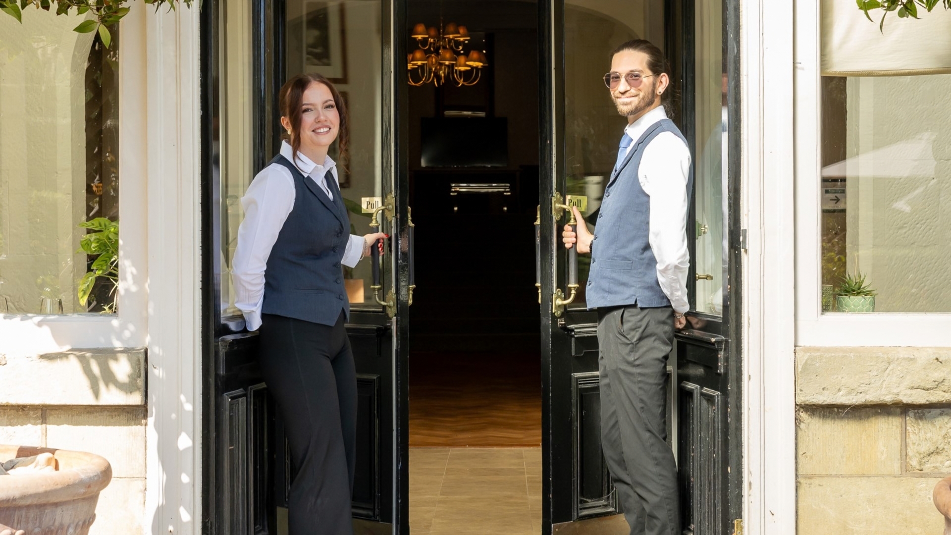 Two hotel team members standing at the entrance of The Royal Hotel, holding open black double doors to welcome guests. The elegant entrance is framed by large windows, potted plants and views into the warmly lit interior.