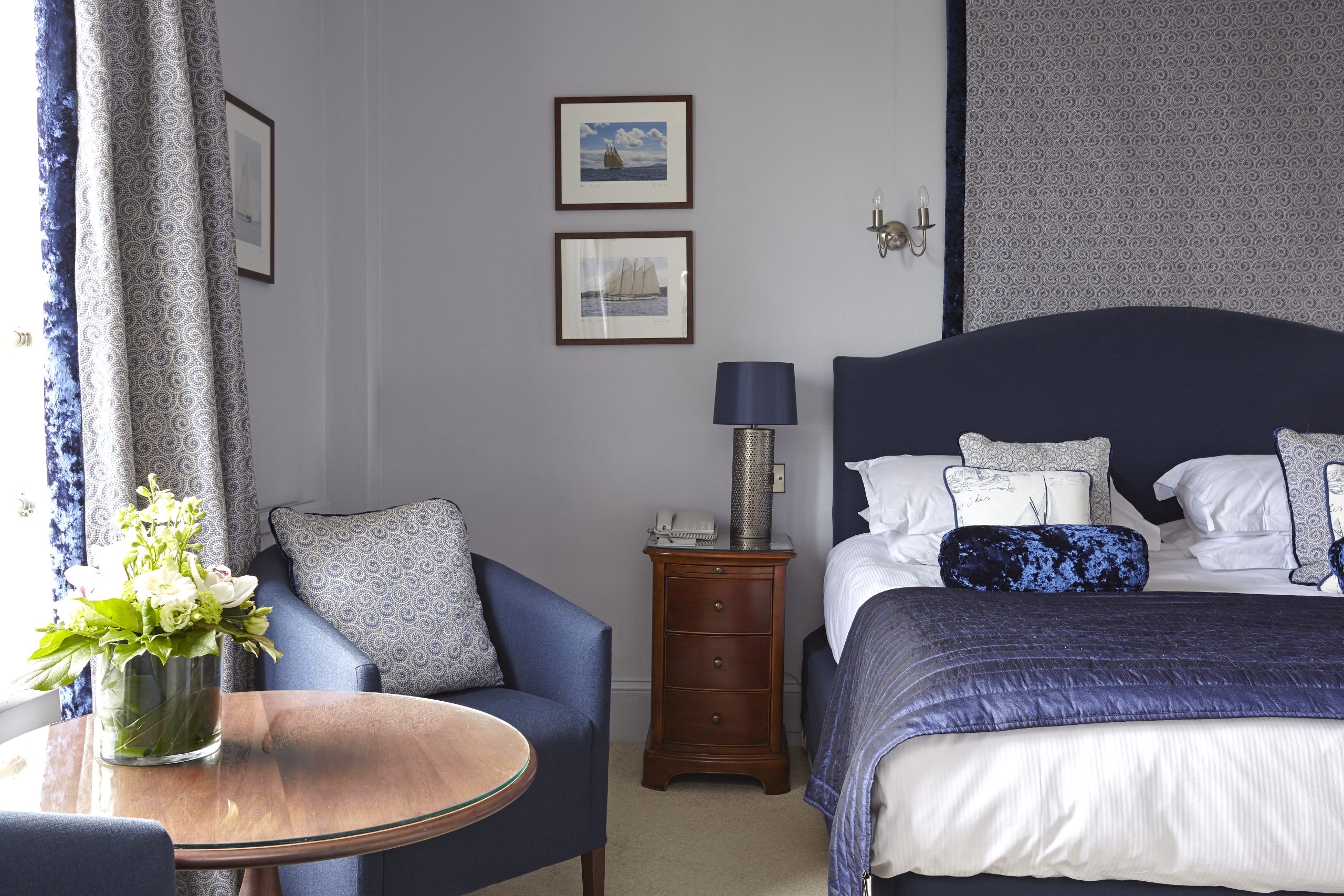 Hotel bedroom with blue furnishings, patterned curtains and a small seating area.