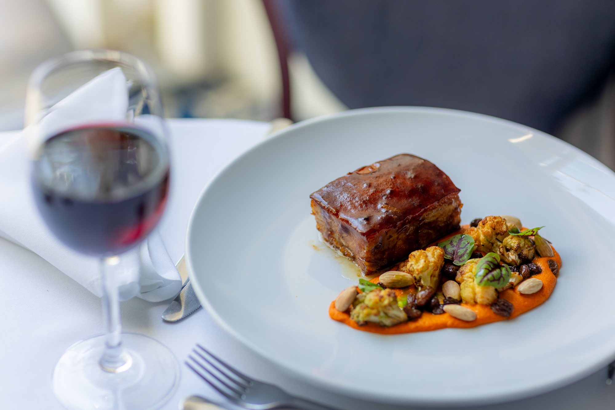 A plated gourmet dish with a glazed rectangular piece of meat served over a bright orange puree, garnished with roasted cauliflower, nuts, and herbs, alongside a glass of red wine on a white table.