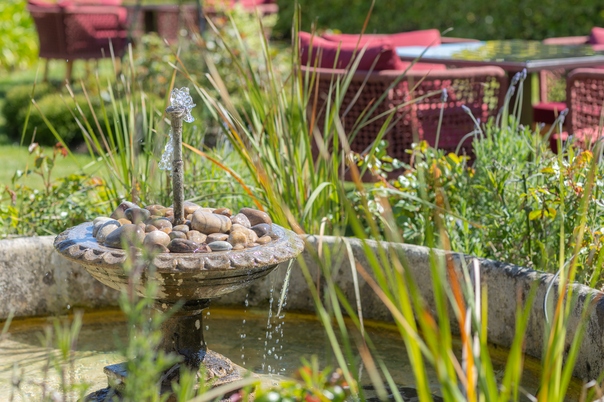 A small garden fountain with water trickling over stones, surrounded by green plants and outdoor seating with red cushions in the background.