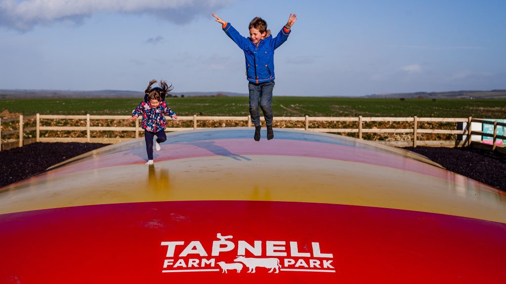 Two children play on a giant outdoor jumping pillow at Tapnell Farm Park, surrounded by countryside views and open fields.