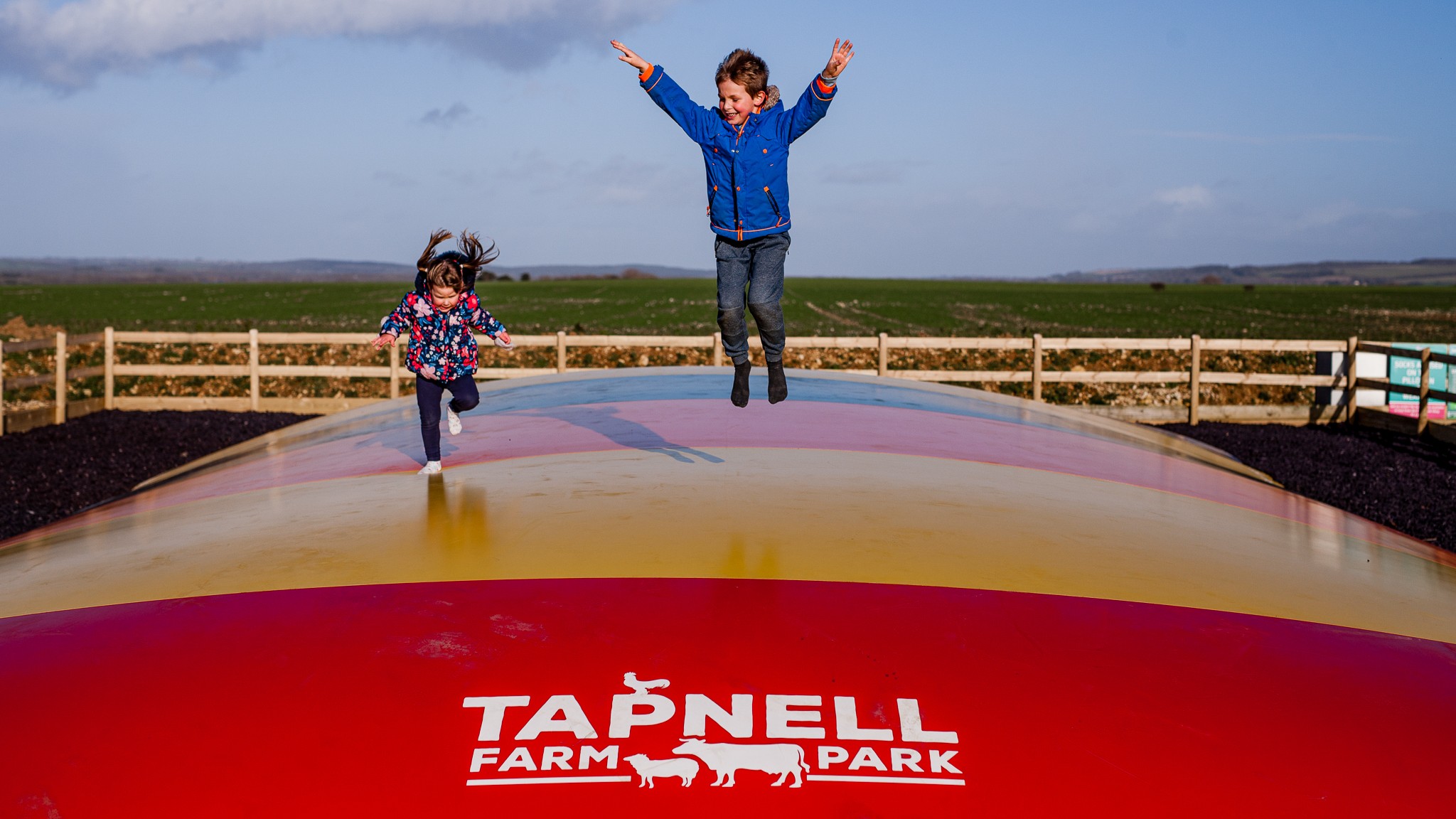 Two children play on a giant outdoor jumping pillow at Tapnell Farm Park, surrounded by countryside views and open fields.