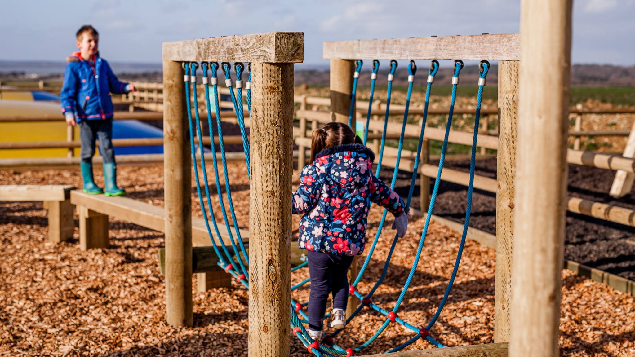 A child climbs across a rope bridge in an outdoor adventure playground at Tapnell Farm, with another child standing nearby.