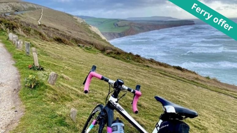 Road bike parked beside a coastal path overlooking dramatic cliffs and the sea on the Isle of Wight. Rolling green hills lead to rugged coastline views under a cloudy sky, with a green corner ribbon reading “Ferry offer”.