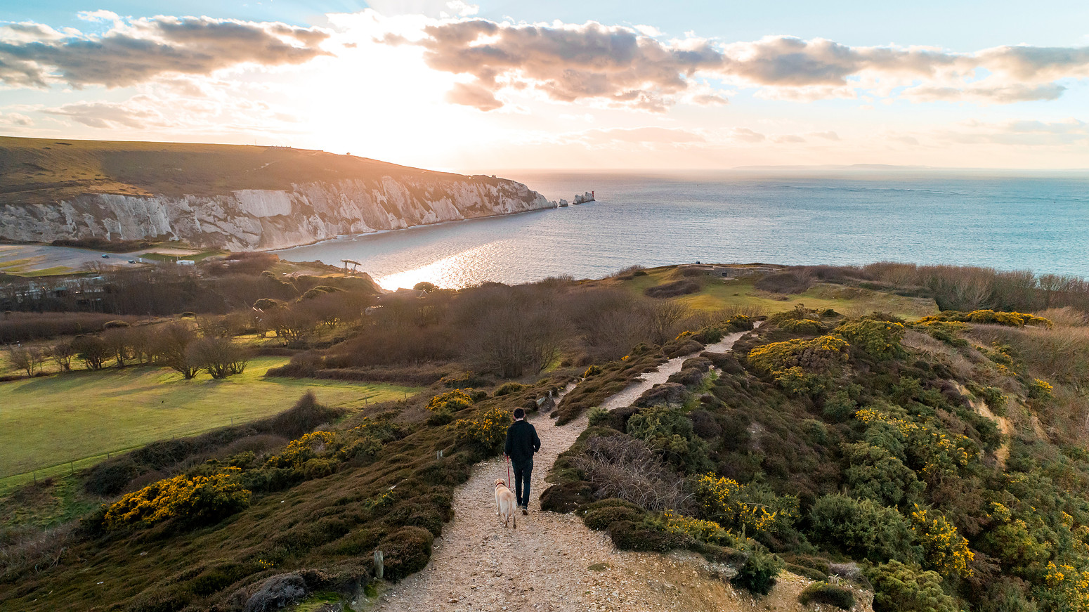 A man walking a dog along a coastal path on Headon Warren with views of the Needles rock formations and the sea at sunset on the Isle of Wight.