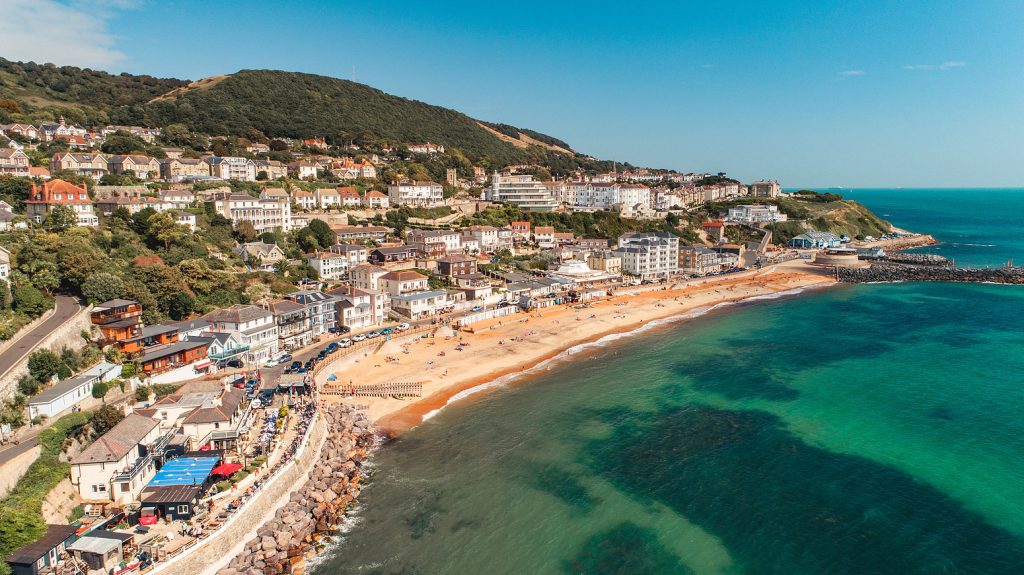 A sunny aerial view of Ventnor on the Isle of Wight, showing colourful houses, sandy beach, and turquoise sea with people relaxing by the water.