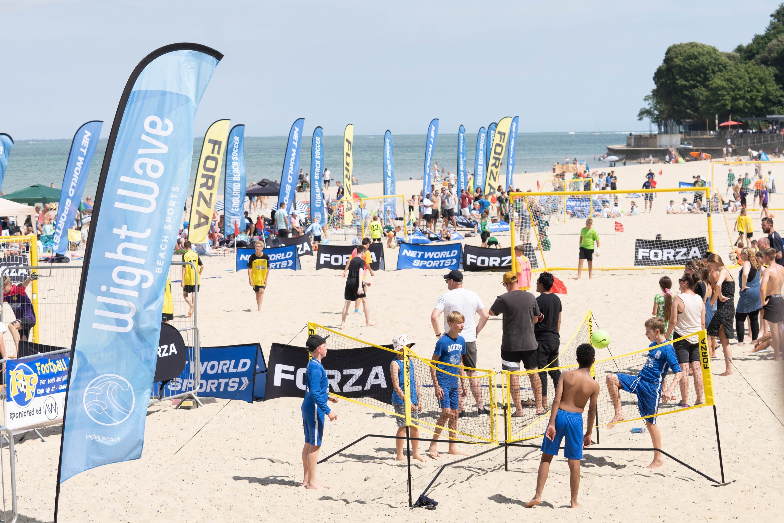 A wide view of a crowded beach soccer event with multiple small soccer goals on the sand. Many children and adults are participating and watching. Tall banners with logos, including “Wight Wave” and “FORZA,” line the playing area. The sea is visible in the background.