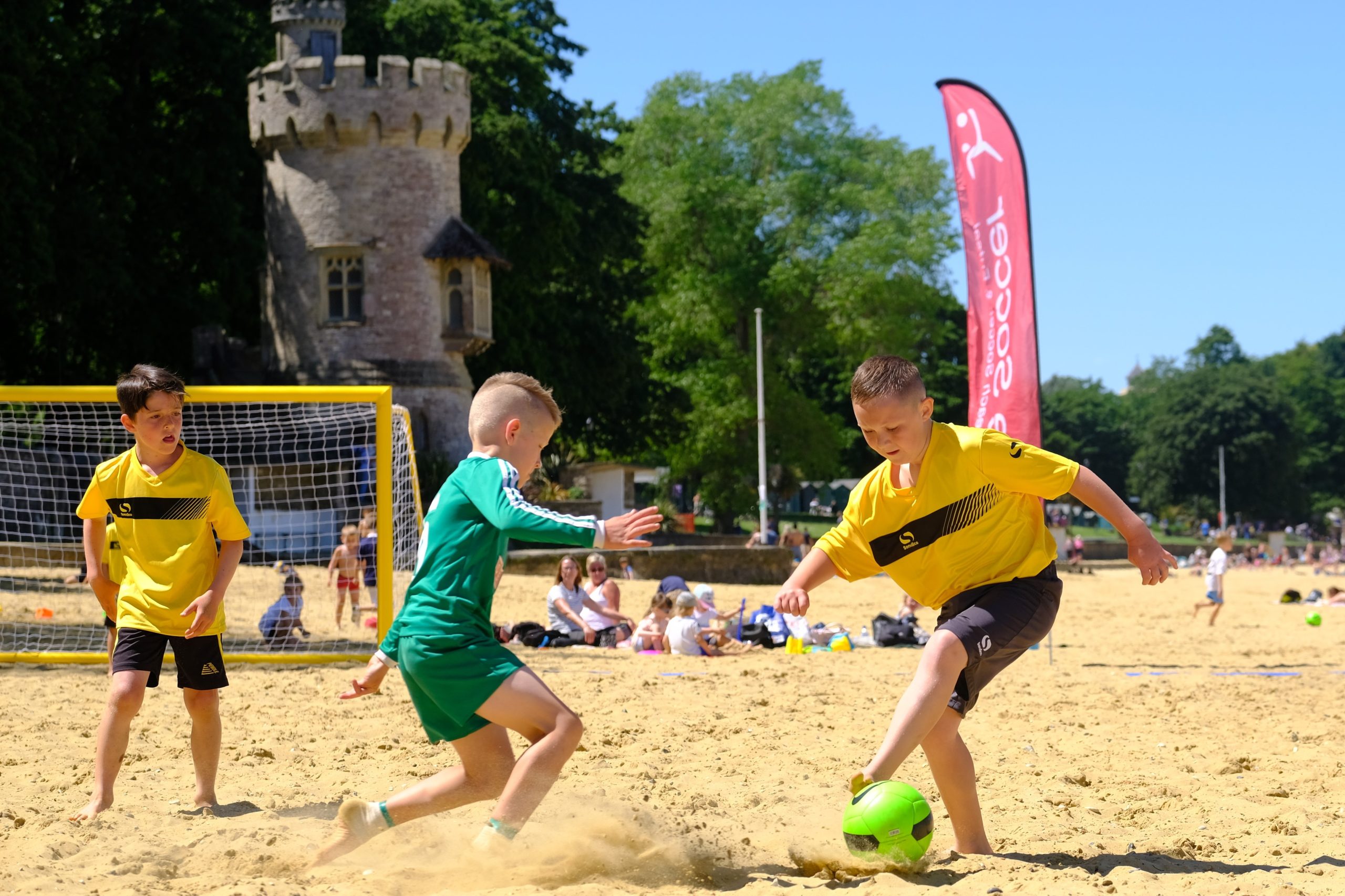 Three children play beach soccer on sand. Two boys in yellow jerseys and one boy in a green jersey are focused on a bright green soccer ball. In the background, there is a small stone castle tower, and a red vertical banner is visible.