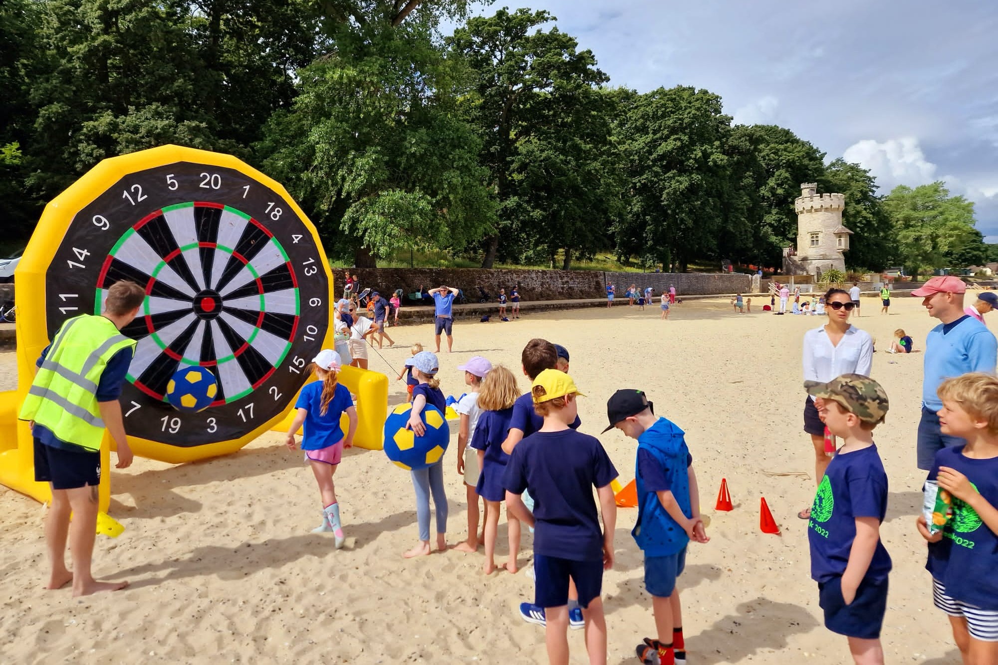 Children participate in an inflatable soccer dart game on the sand, kicking large soft soccer balls toward a giant dartboard. Adults and other kids watch nearby. A stone tower is visible in the background.