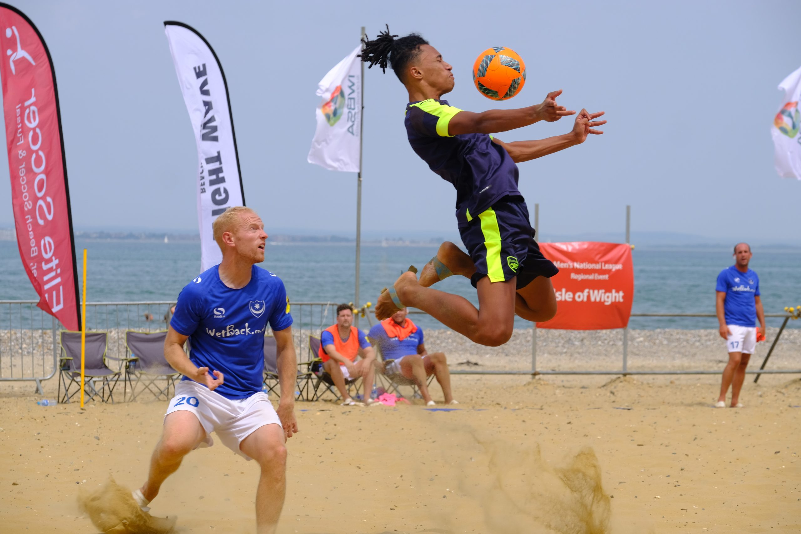 Two male players compete in beach soccer on sand. A player in a dark blue and neon uniform jumps mid-air to chest the orange ball, while a player in a blue jersey with the number 20 watches closely.