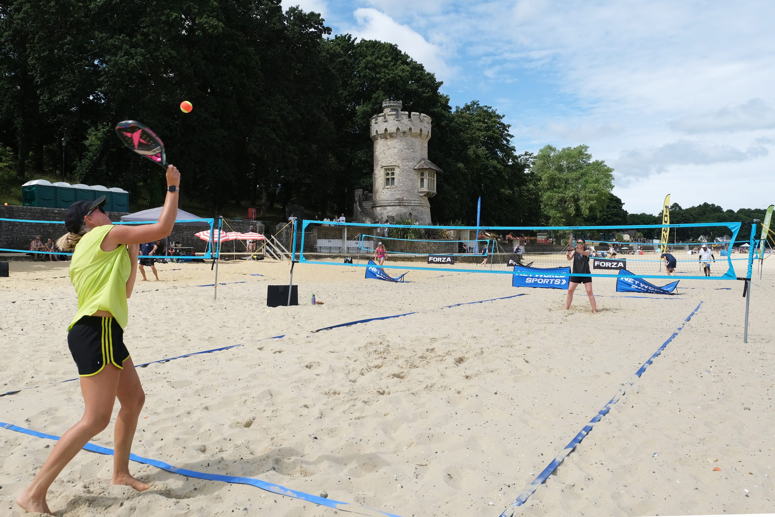 A woman in a yellow top and black shorts is preparing to serve or hit a ball in a beach pickleball or paddle court. Blue nets divide multiple courts on the sand, with a stone tower visible in the background and spectators watching.
