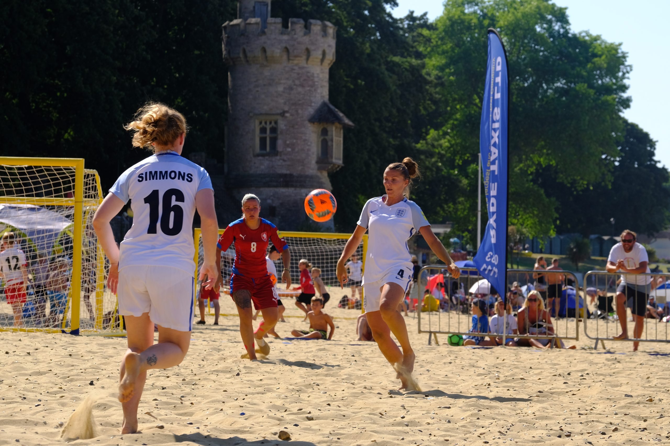 Three female players are engaged in a beach soccer match on sand. Two players in white jerseys, one with the name “Simmons” and number 16, are focused on an orange ball mid-air. A player in a red jersey with number 8 stands in goal. A small stone tower is in the background, and a blue vertical banner is nearby.