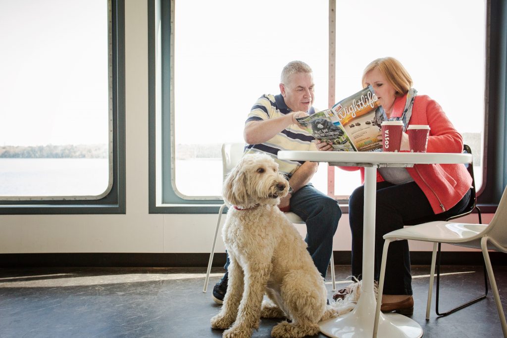 A couple with a dog onboard a ferry next to windows, with coffee and a magazine
