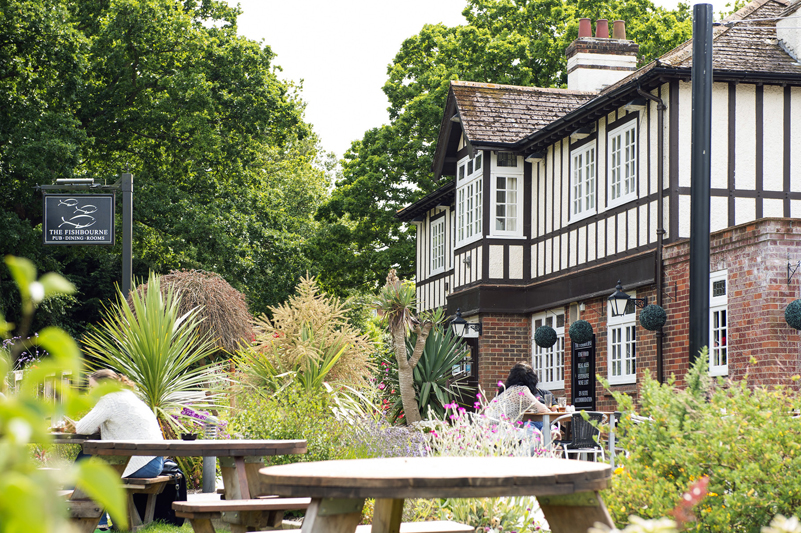 The outside of a pub with a beer garden, tables and trees in the foreground. The pub has a mock-Tudor style exterior