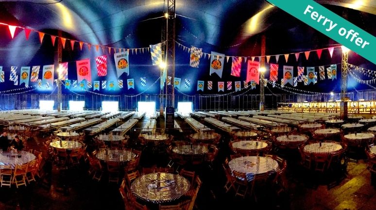 Interior of a large Oktoberfest-style beer tent set up with long wooden banquet tables and round tables covered in blue-and-white checked cloths.