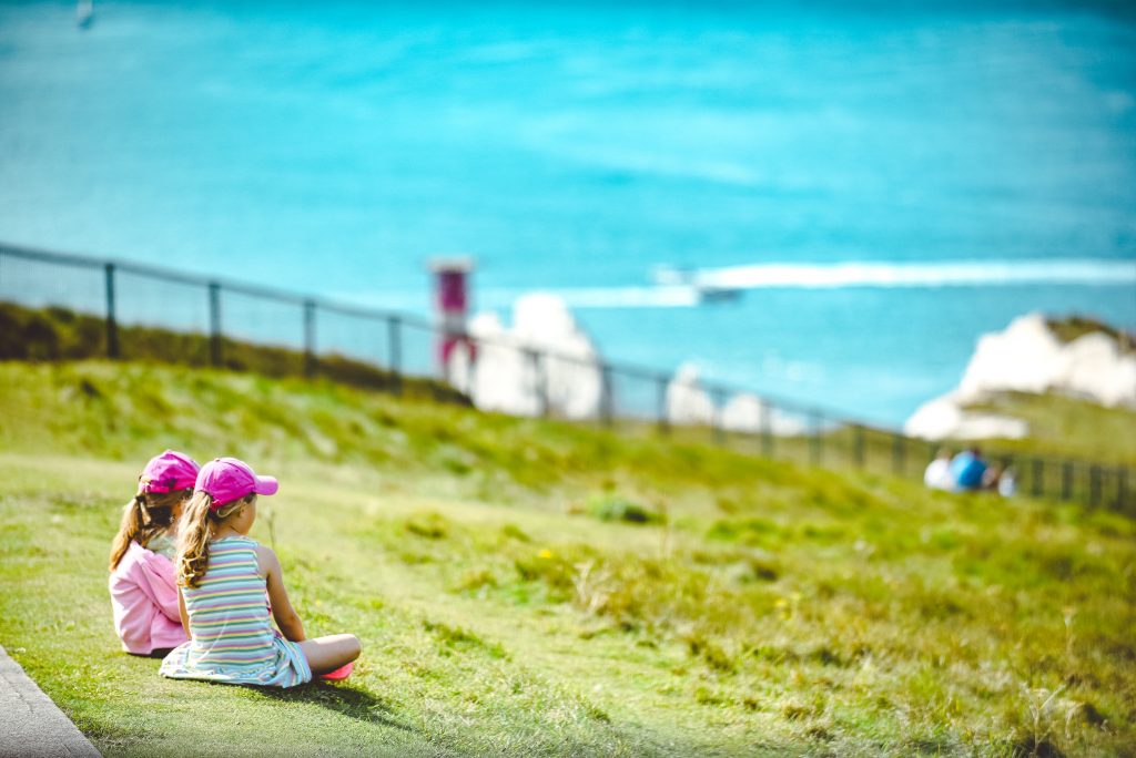 Two young girls sitting on a grassy hillside overlooking The Needles rock formation on The Isle of Wight