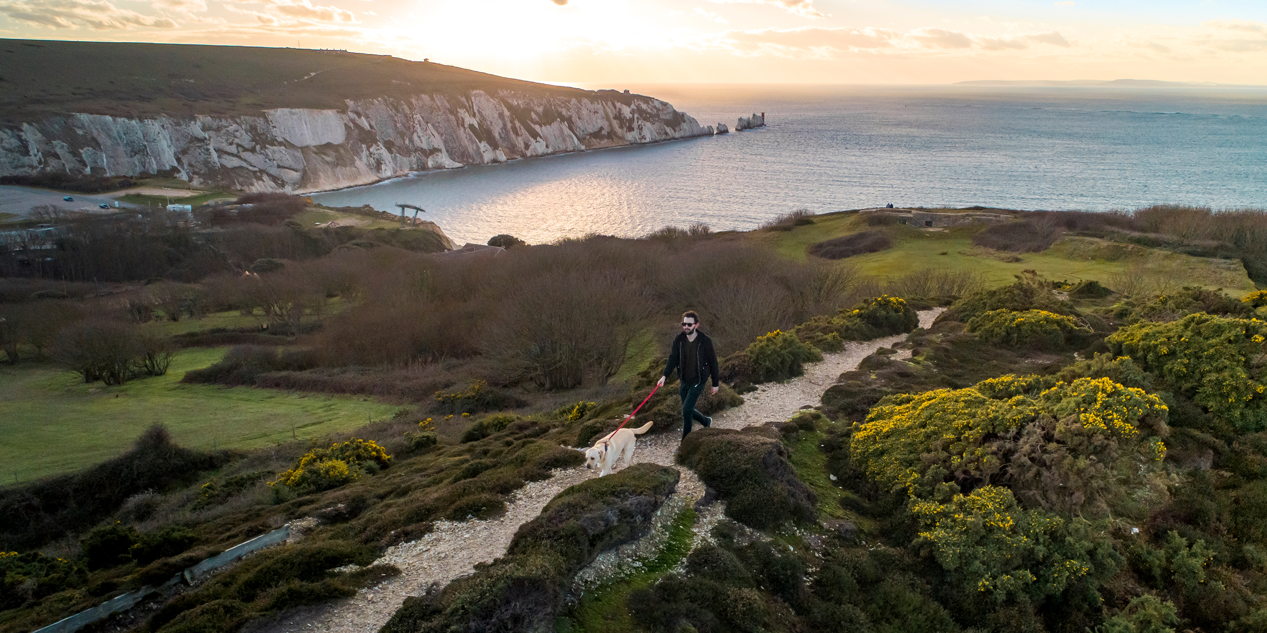 A man walking a dog across a moor overlooking the sea and the Needles on the Isle of Wight