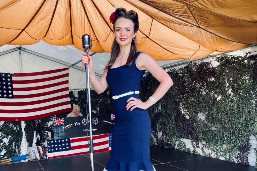 Vintage performer on an E.N.S.A stage, wearing a 1940s-style outfit and holding a microphone, with wartime flags and camouflage netting in the background.