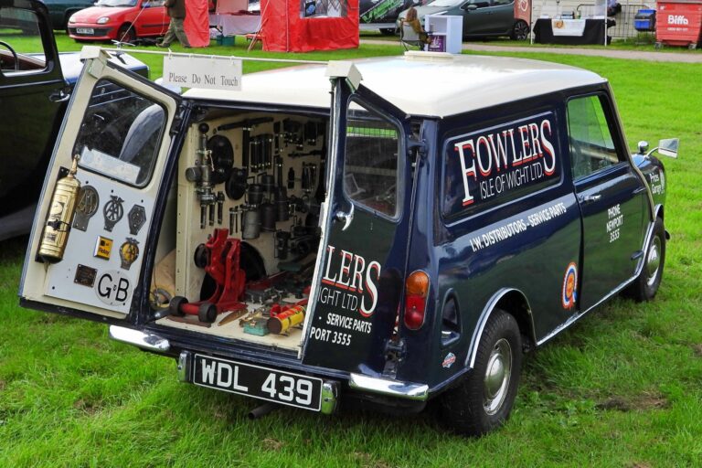 A blue and white vintage Mini van with the words "Fowlers Isle of Wight Ltd." on the side. The back door is open, revealing an assortment of tools and mechanical parts inside the cargo area, with a sign saying "Please Do Not Touch.