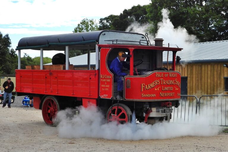 A red, steam-powered vintage truck with a black roof, displaying "Isle of Wight Farmers Trading Co." on the side. A person dressed in blue is driving the truck, while smoke is billowing out from the engine