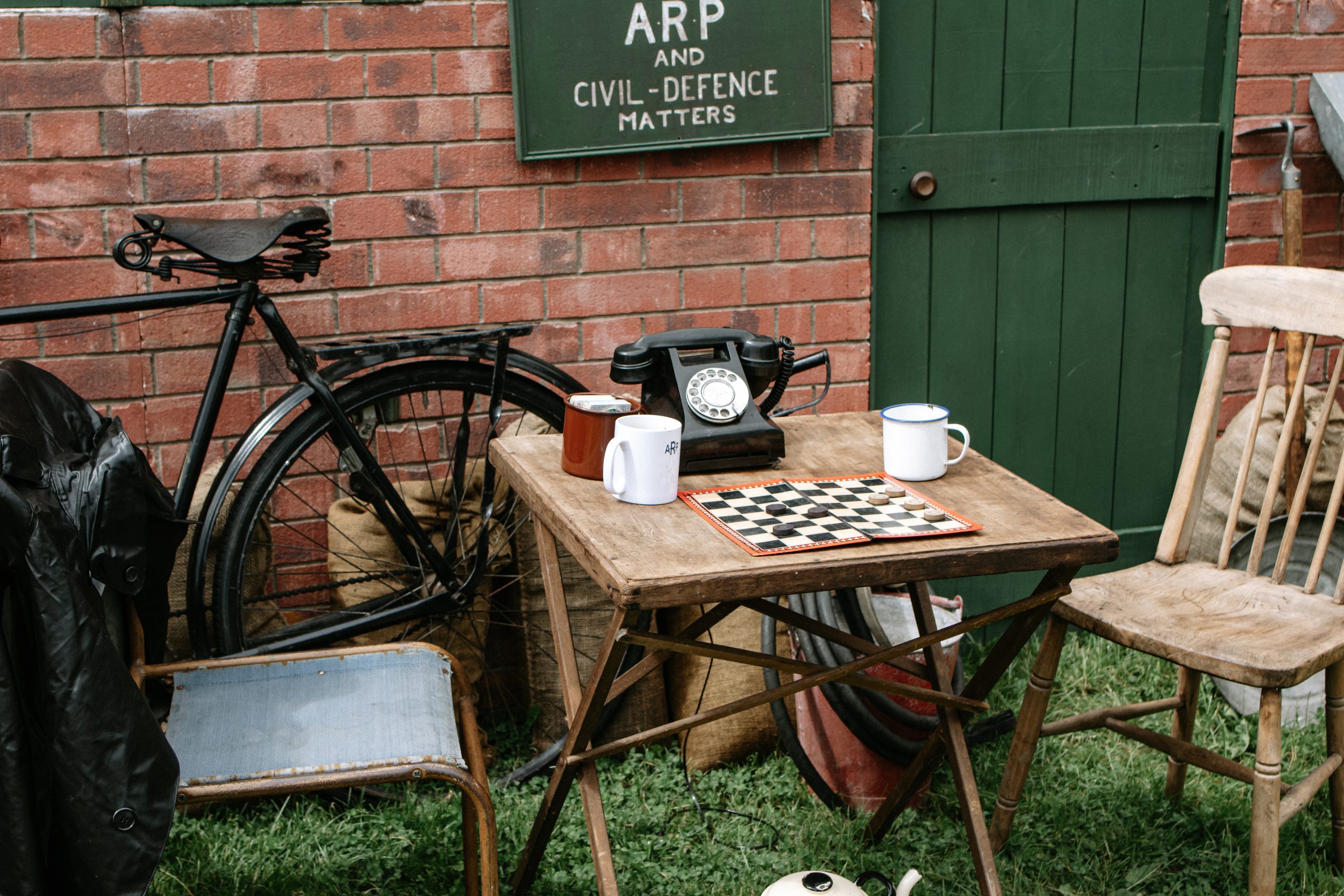 1940s street scene with a wooden table set for tea, a chessboard, enamel mugs, a rotary telephone and a bicycle beside a brick wall with wartime signage.