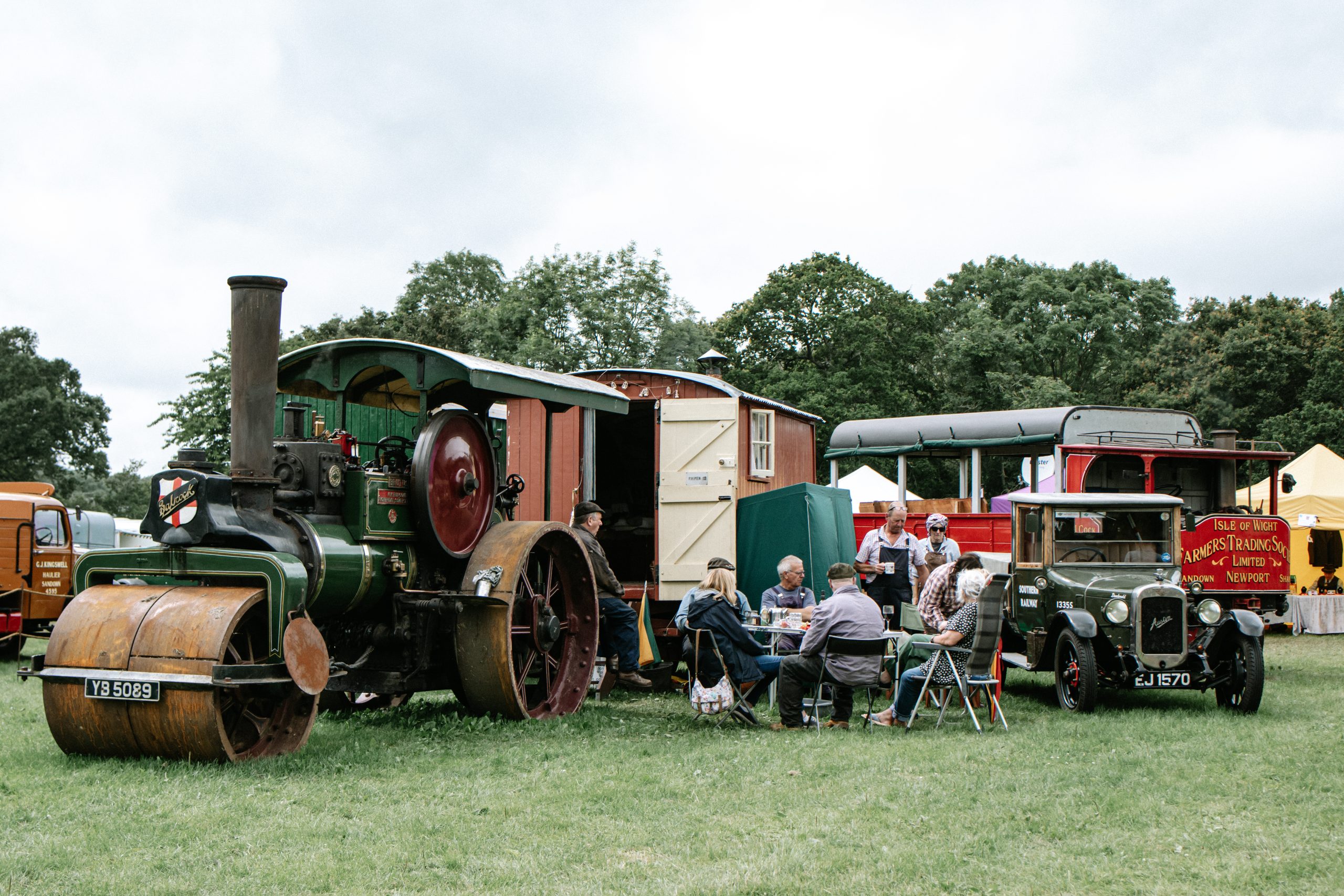 Visitors seated outdoors among historic steam machinery and vintage vehicles at a 1940s-themed event, set on a grassy field with trees in the background.
