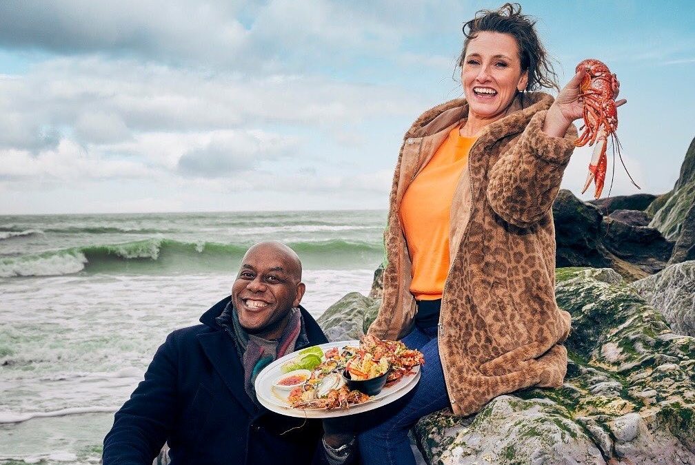Ainsley Harriott and Grace Dent on the rocks of Ventnor beach holding seafood