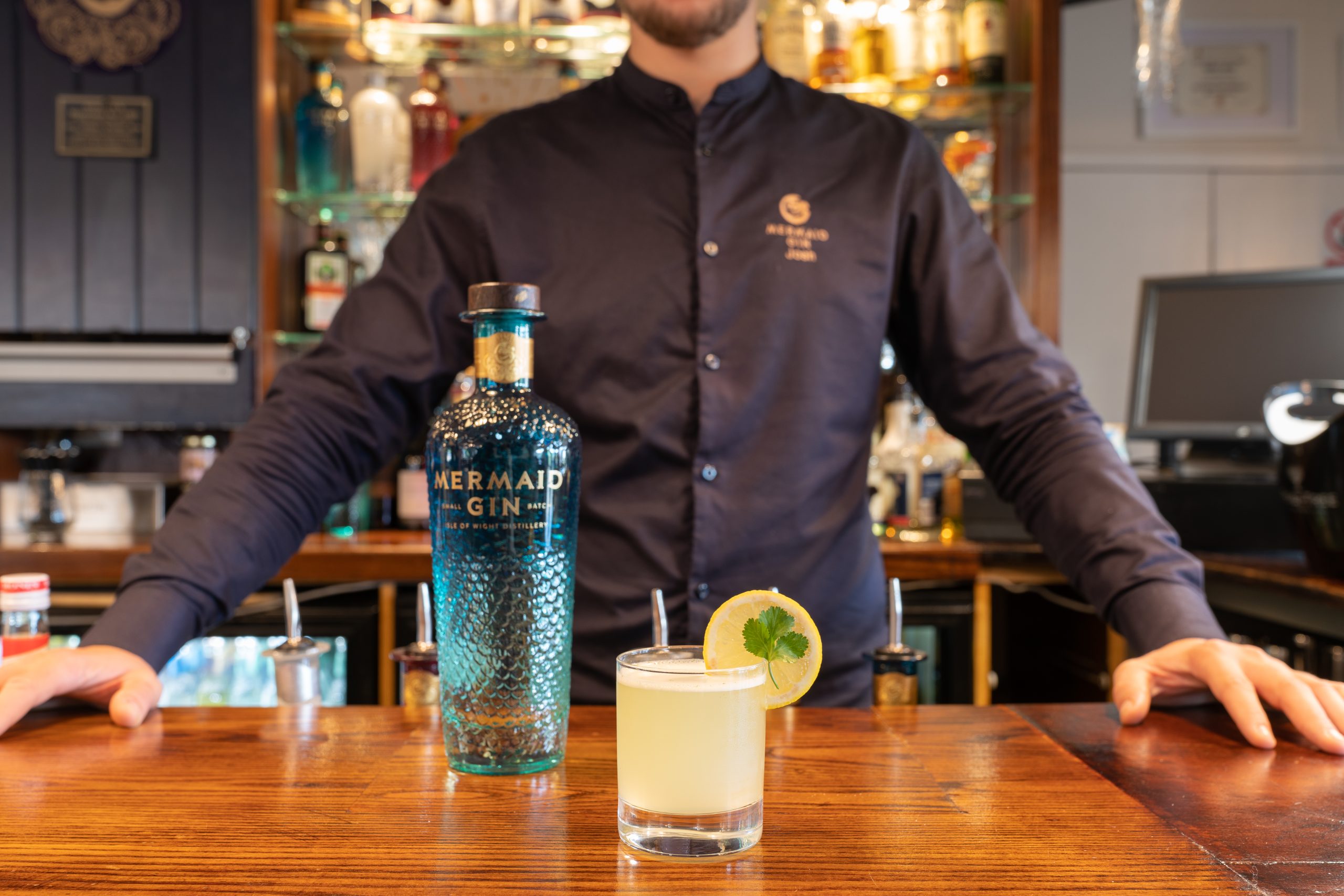 A wooden bar with a bottle of gin and a cocktail on top with a bartender behind - Mermaid Distillery Isle of Wight