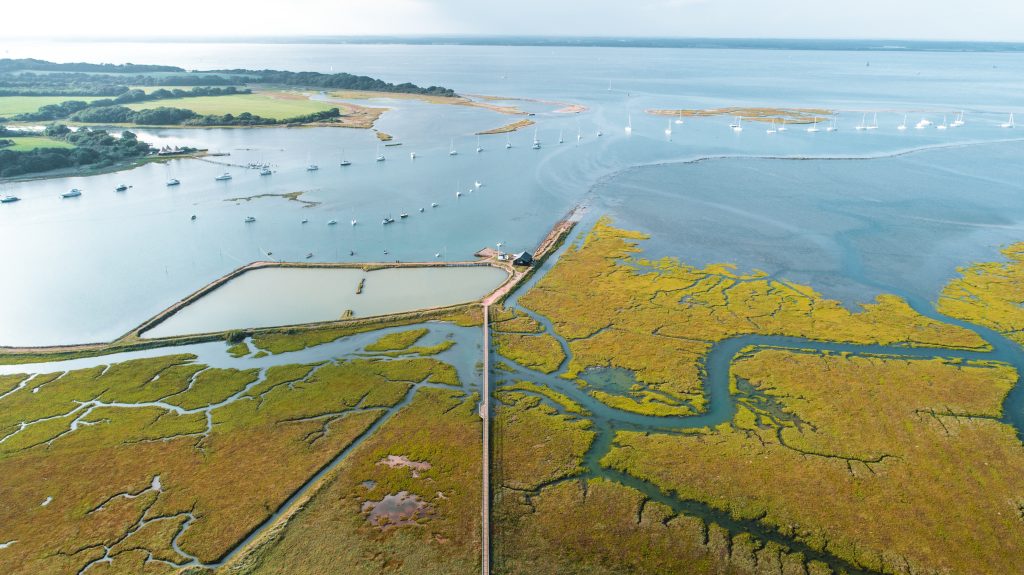 Aerial view of Newtown Creek on the Isle of Wight, showing winding tidal channels, saltmarsh, and moored sailboats in the distance.