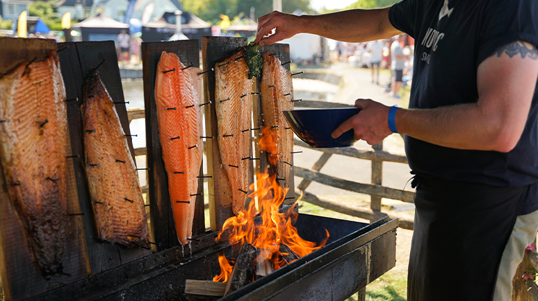 A white male grilling fish on an open fire grill. The fish are pinned to wooden boards on the back of the fire grill, and the man is holding a bowl ready to pick up the fish when it's cooked.