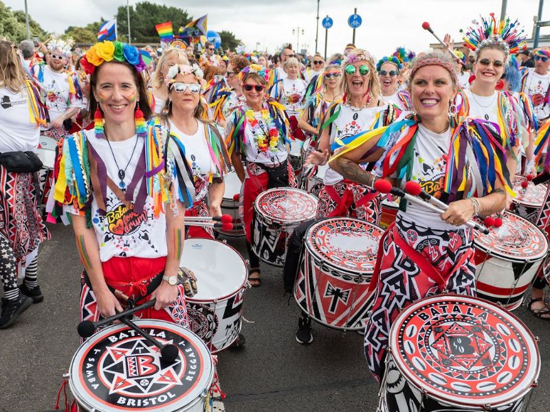 Parade drummers in colourful outfits and rainbow accessories marching along a street.