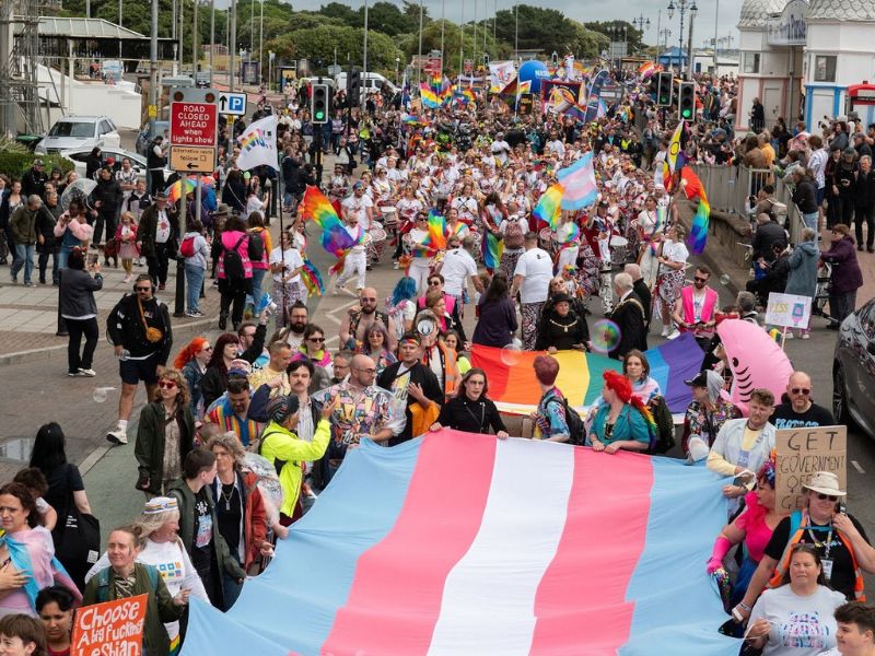 Crowd at a Pride parade carrying a large transgender flag with rainbow flags throughout.