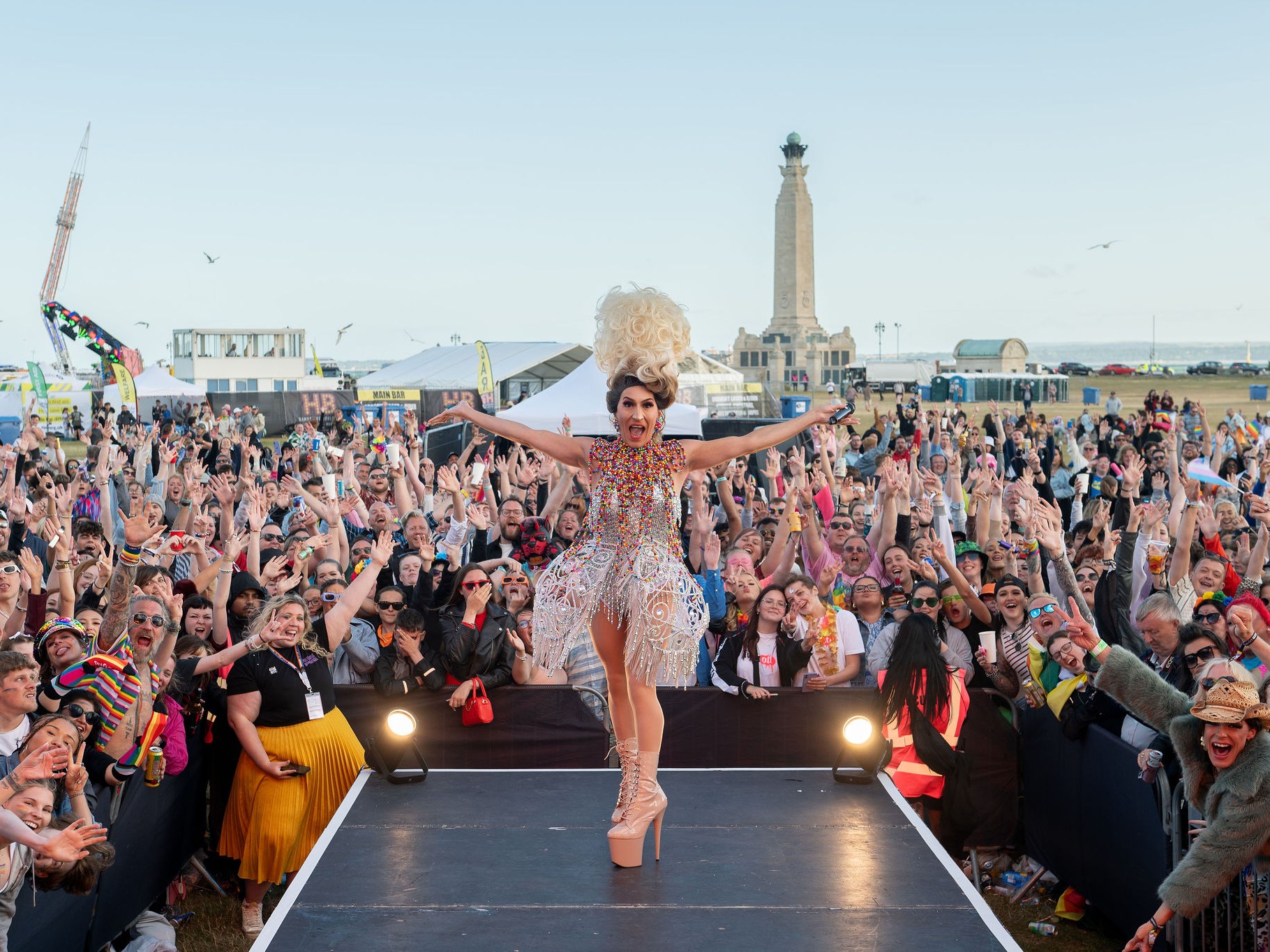 A performer stands on an outdoor stage at Portsmouth Pride, facing a large crowd on Southsea Common, with festival stalls and the Spinnaker Tower visible in the background.