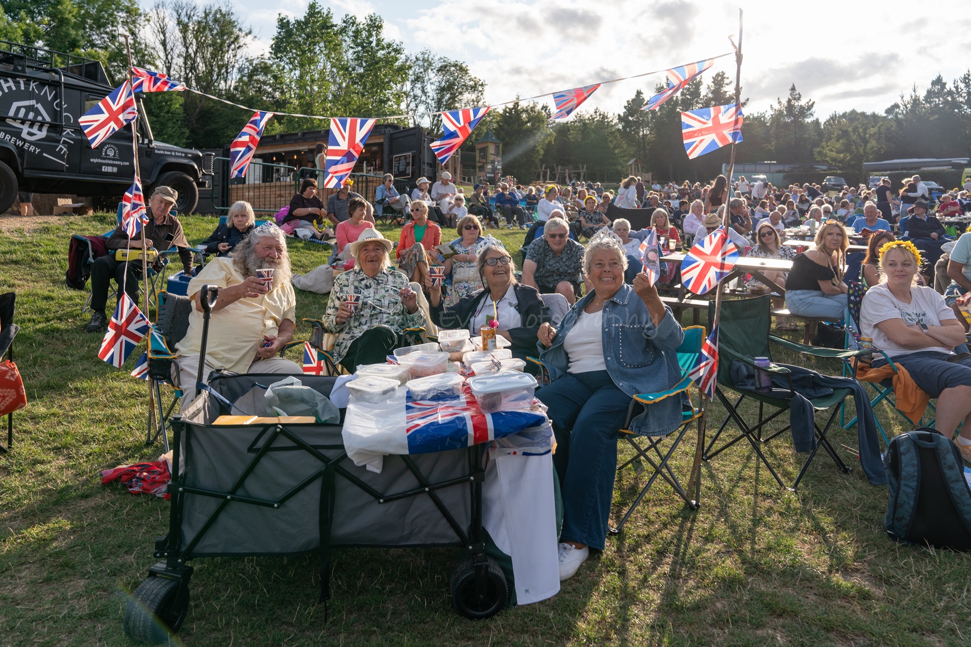 Proms audience relaxing on picnic chairs and benches, waving Union Jack flags during an outdoor evening concert