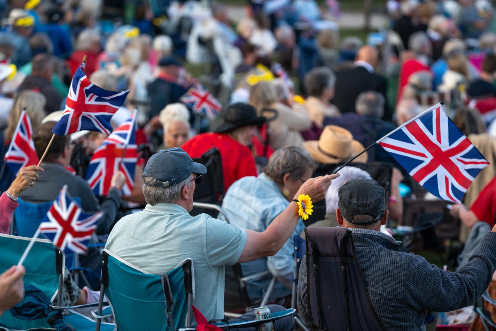 Outdoor Proms audience seated on picnic chairs, waving Union Jack flags across a grassy park setting