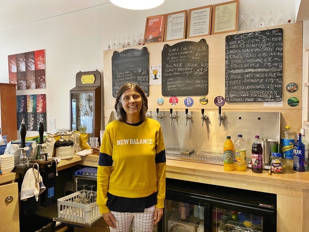 A female in a yellow t-shirt standing behind a bar in front of beers on tap and chalkboard signage