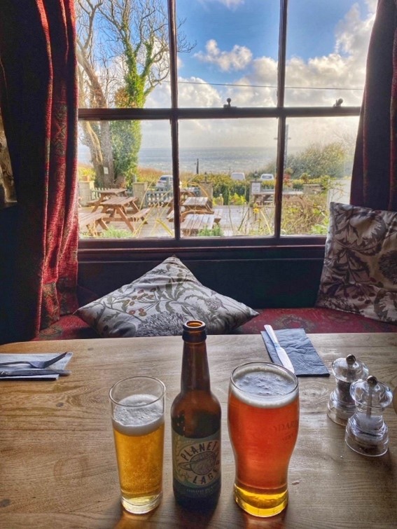 A table inside a pub with 3 beers on it, looking out to a window seat with cushions, and views down to the sea