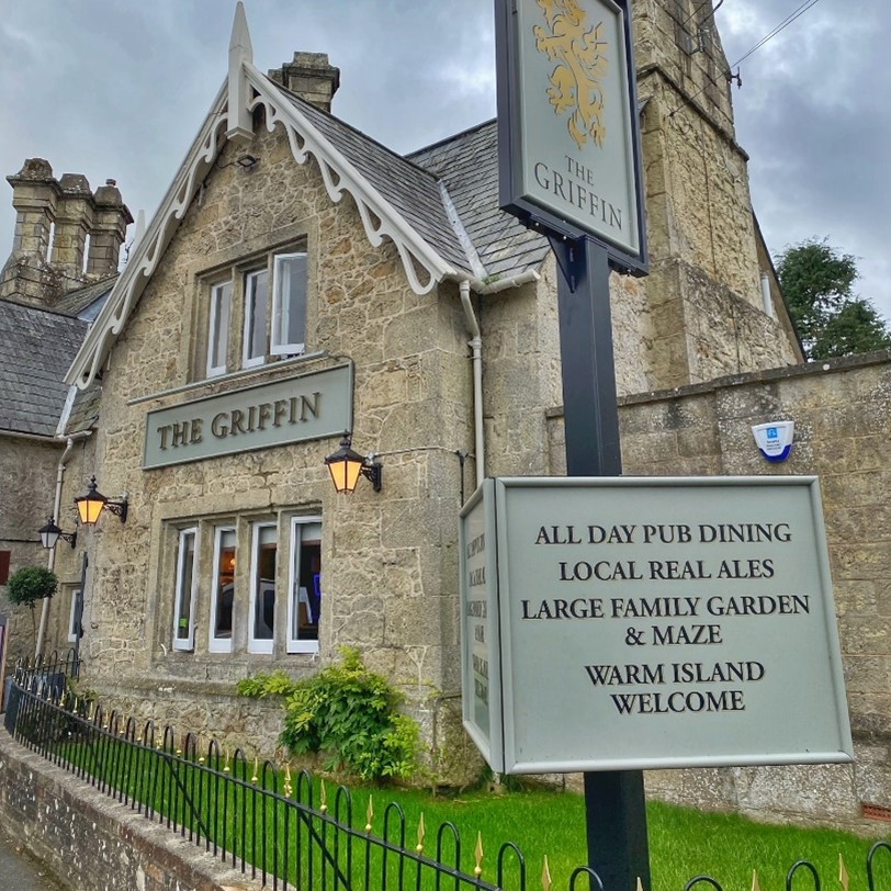 The exterior of a pub, with signage in the foreground, a lawn and sandstone coloured walls