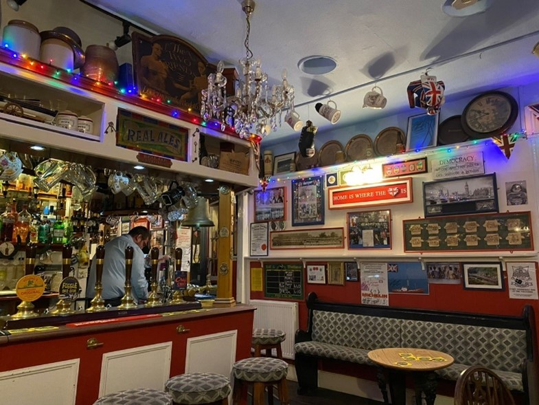 The interior of a small traditional British pub, with bar, bar stools, beer pumps, glasses hanging and memorabilia on the walls