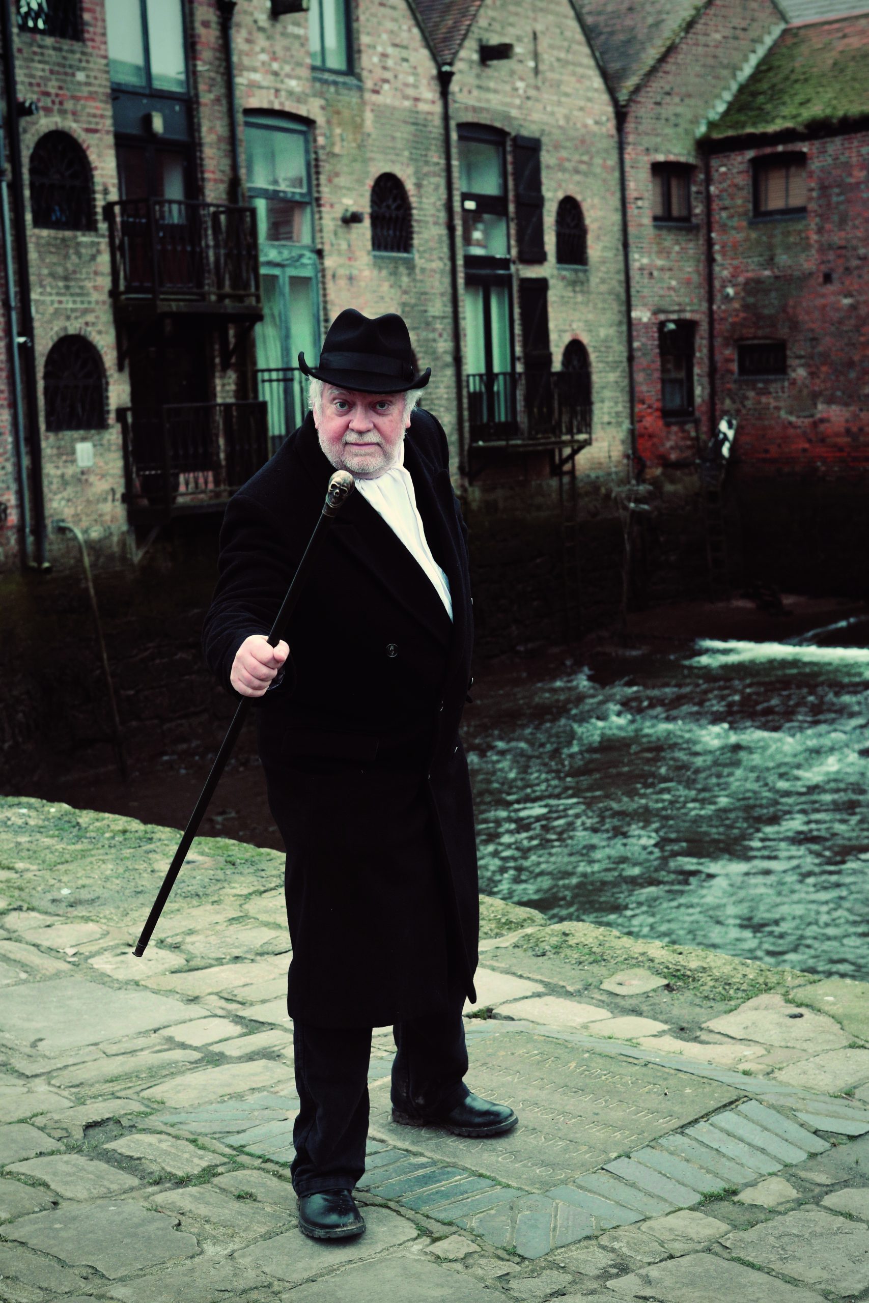 A man standing by a river wearing a black suit and hat, with a walking stick