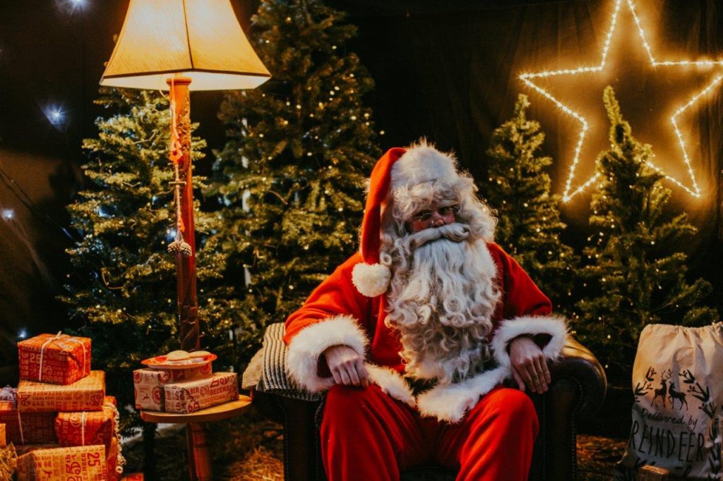 A traditional father Christmas figure sits indoors next to Christmas trees, surrounded by cosy lights and wrapped gifts