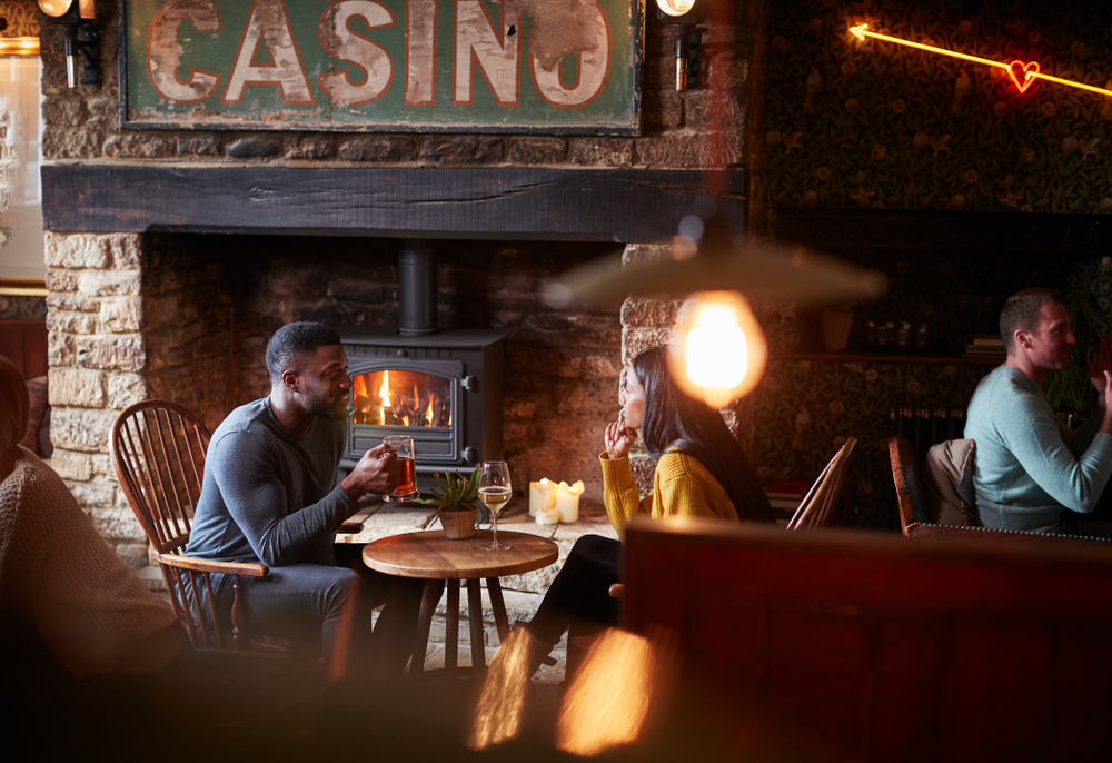 A couple sitting down in front of a log burner, in a cosy looking pub./ There's a glass of wine on the table and the man is holding a pint glass