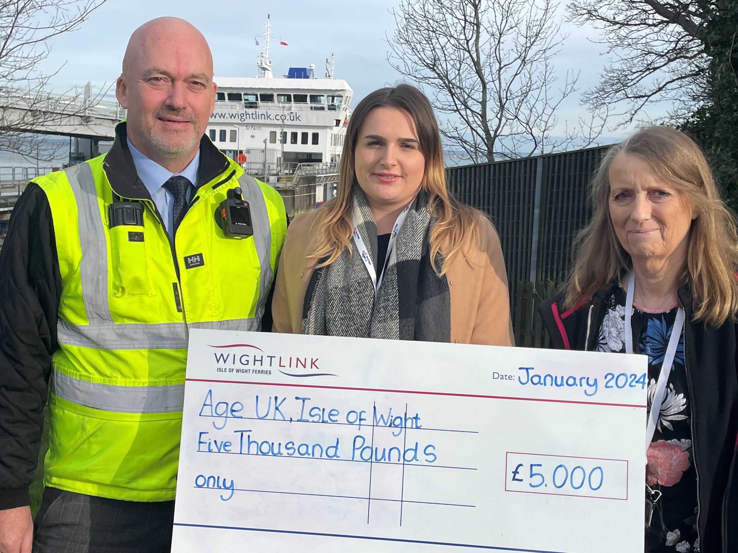 A male Wightlink worker in a high visibility jacket hands a giant cheque to two women, in front of a Wightlink ferry