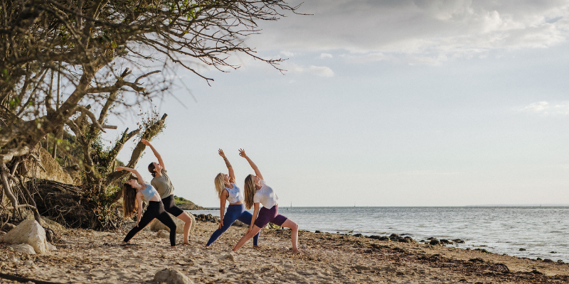 A group of 4 women in sportswear doing yoga poses on a beach on the Isle of Wight