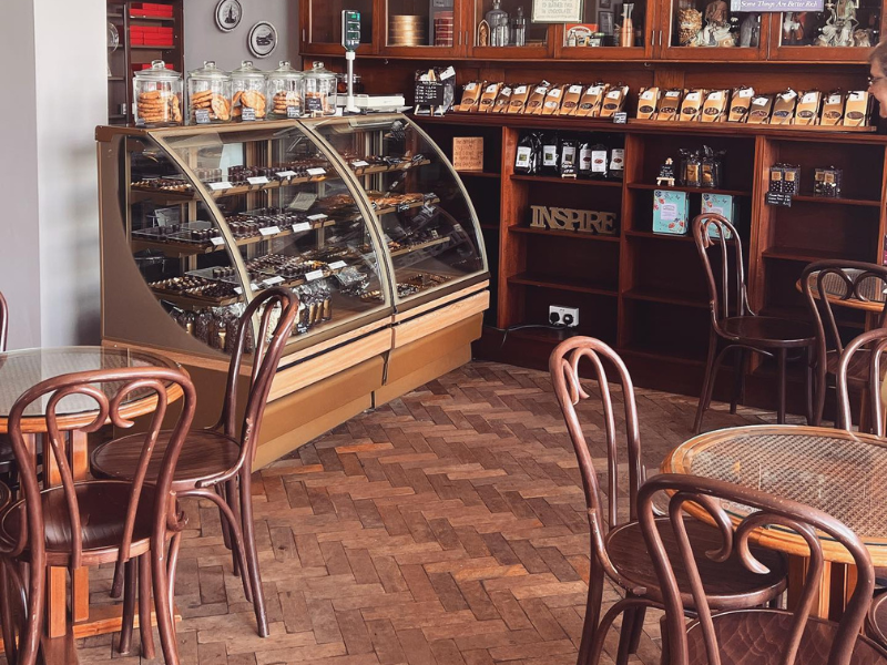 A cafe with parque flooring, whicker furniture and a glass counter full of chocolate